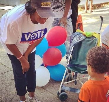 Woman in a 'VOTE' shirt talking to a child near colorful balloons and a stroller.