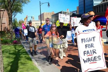 Protesters marching with signs demanding fair wages on a sunny city street.