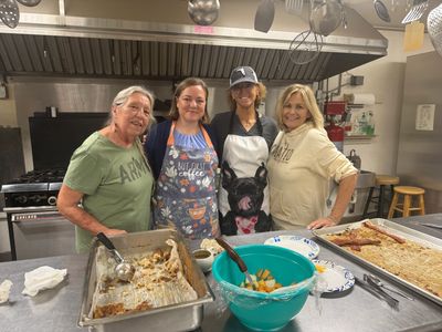 Four women standing in a kitchen with food trays and a salad bowl.