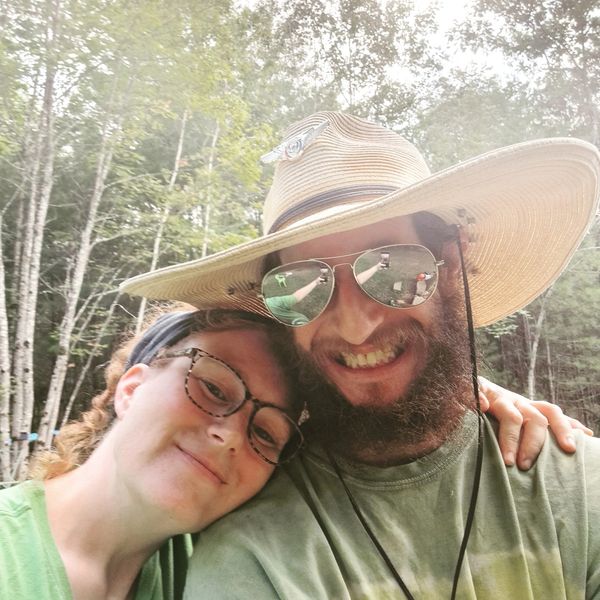 Smiling couple outdoors with man in large hat and sunglasses, woman resting head on his shoulder.