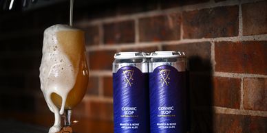 Foamy craft beer poured in a glass beside two cans against a brick wall.