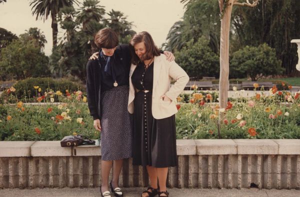 Two women pose together in front of a low wall and flowers.