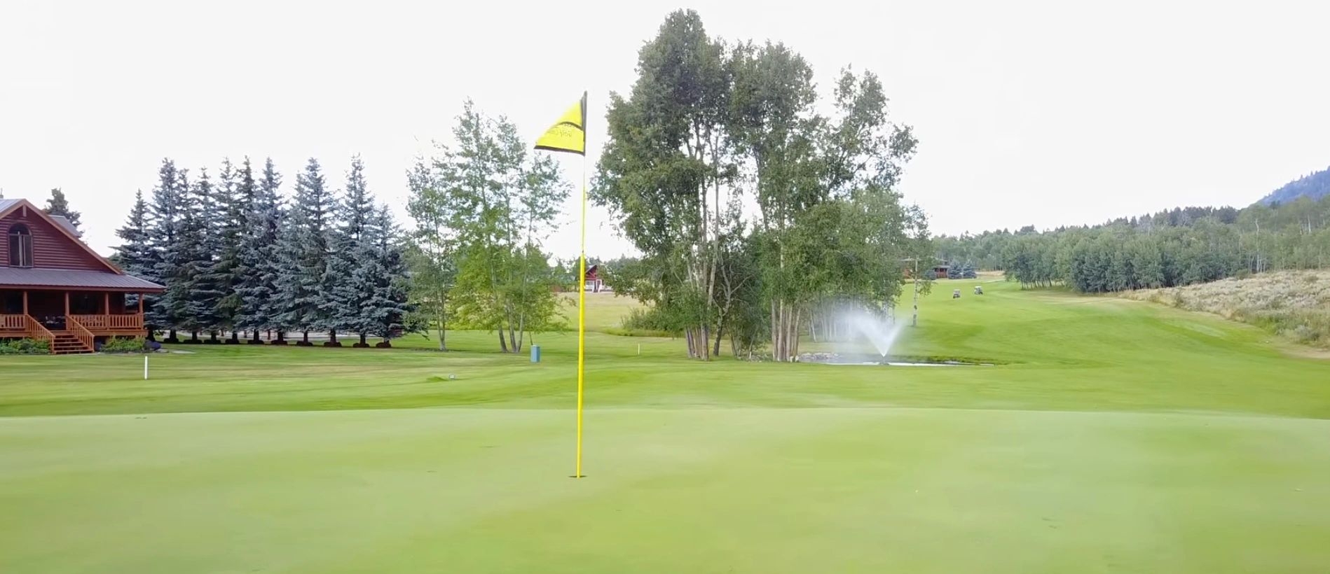 A golf course green with a yellow flag, trees, and a water fountain in the background.