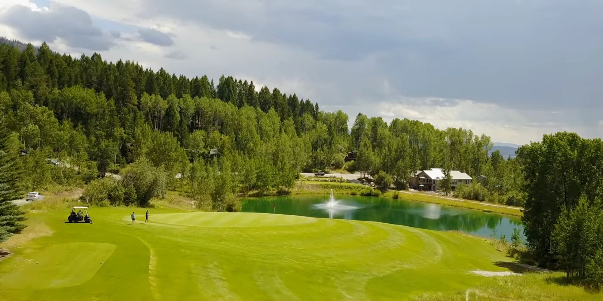 Golfers on a green near a pond with a fountain surrounded by trees.