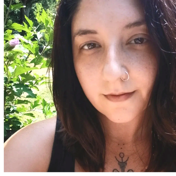Close-up of a woman with a nose ring and tattoo, surrounded by greenery.