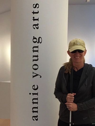 annie, wearing a hat, stands next to a pillar smiling that vertically reads annie young arts 
