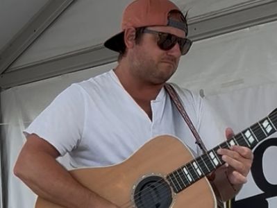 Man wearing sunglasses and cap playing an acoustic guitar.