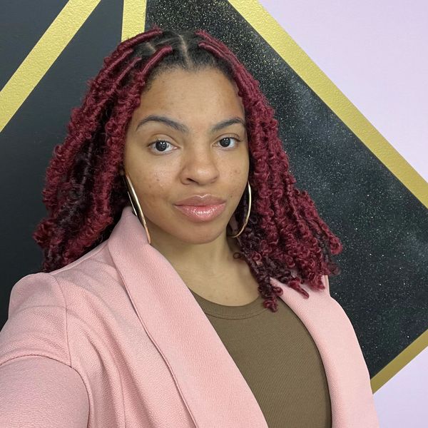 Woman with red curly hair wearing a pink blazer and hoop earrings.