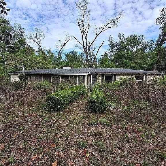 An old, abandoned house surrounded by overgrown bushes and trees under a cloudy sky.