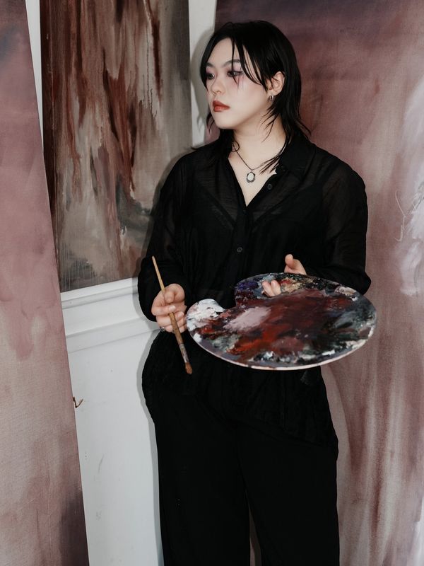 Artist in black holding a paint palette and brush, standing near abstract art.
