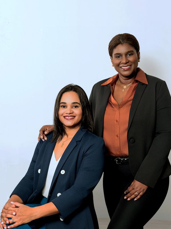 Two professional women smiling, one seated and one standing, dressed in business attire.