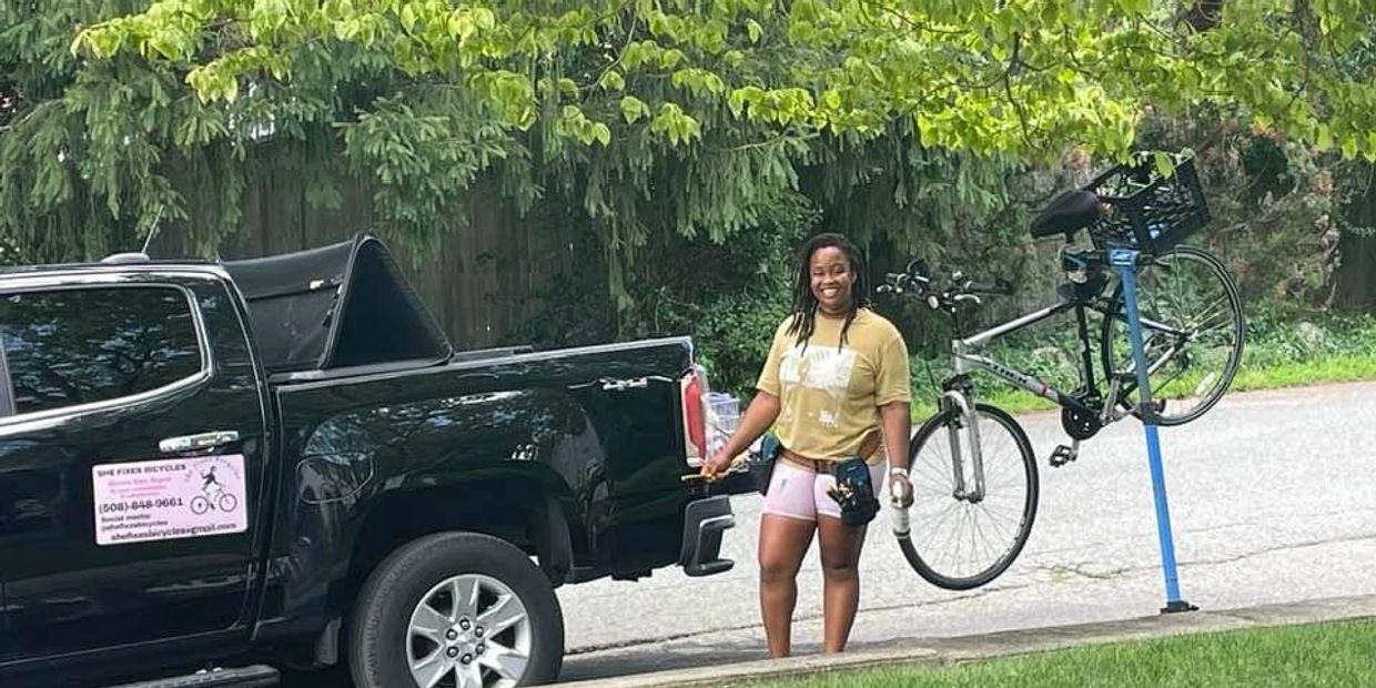 A woman stands next to a black pickup and a bike repair stand with a bike.