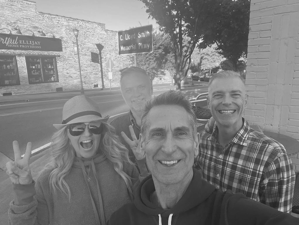 Four adults smiling and posing for a selfie on a sunny street.