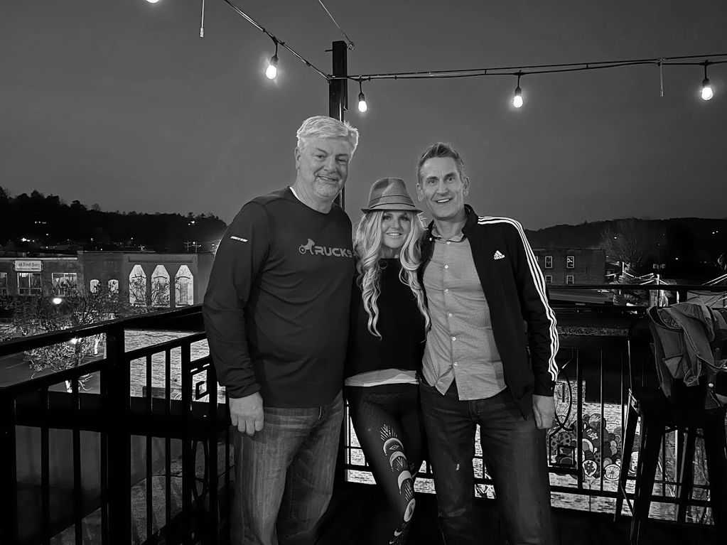 Three friends posing happily on a balcony at night under string lights.