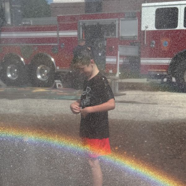 Boy stands in sprinkler water with a rainbow and fire trucks behind.