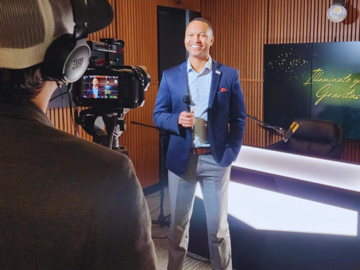 Man in blue blazer holding microphone being filmed in studio.