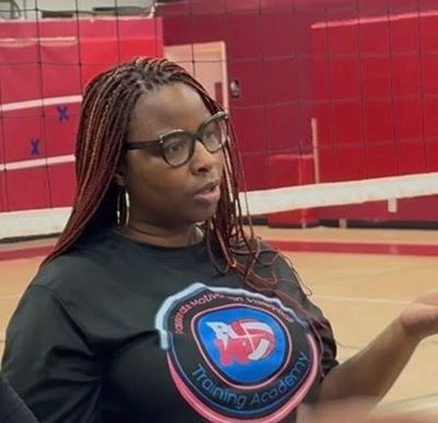 Woman with braided hair and glasses speaking in a gym with a volleyball net.