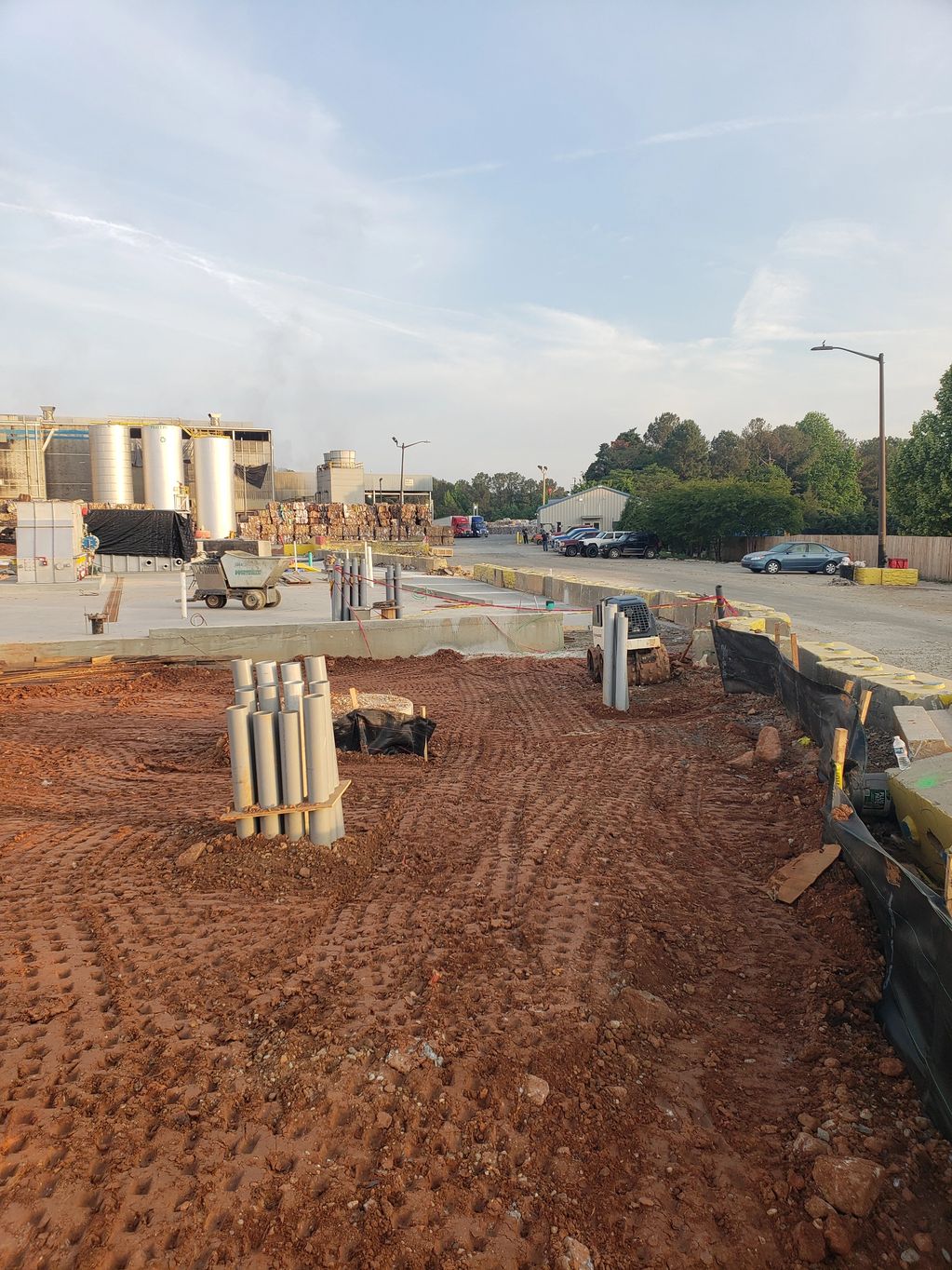 Construction site with exposed pipes and red soil near industrial buildings.