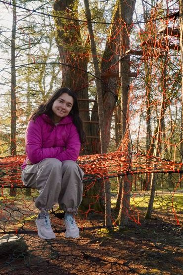 A young woman sits on an orange rope net structure in a forested park.