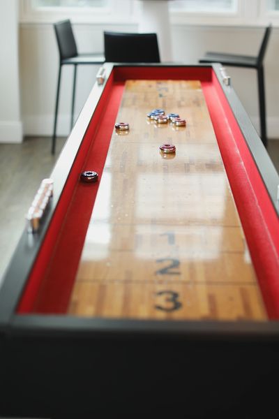 A wooden shuffleboard table with red rails and several pucks, two black chairs and a table.