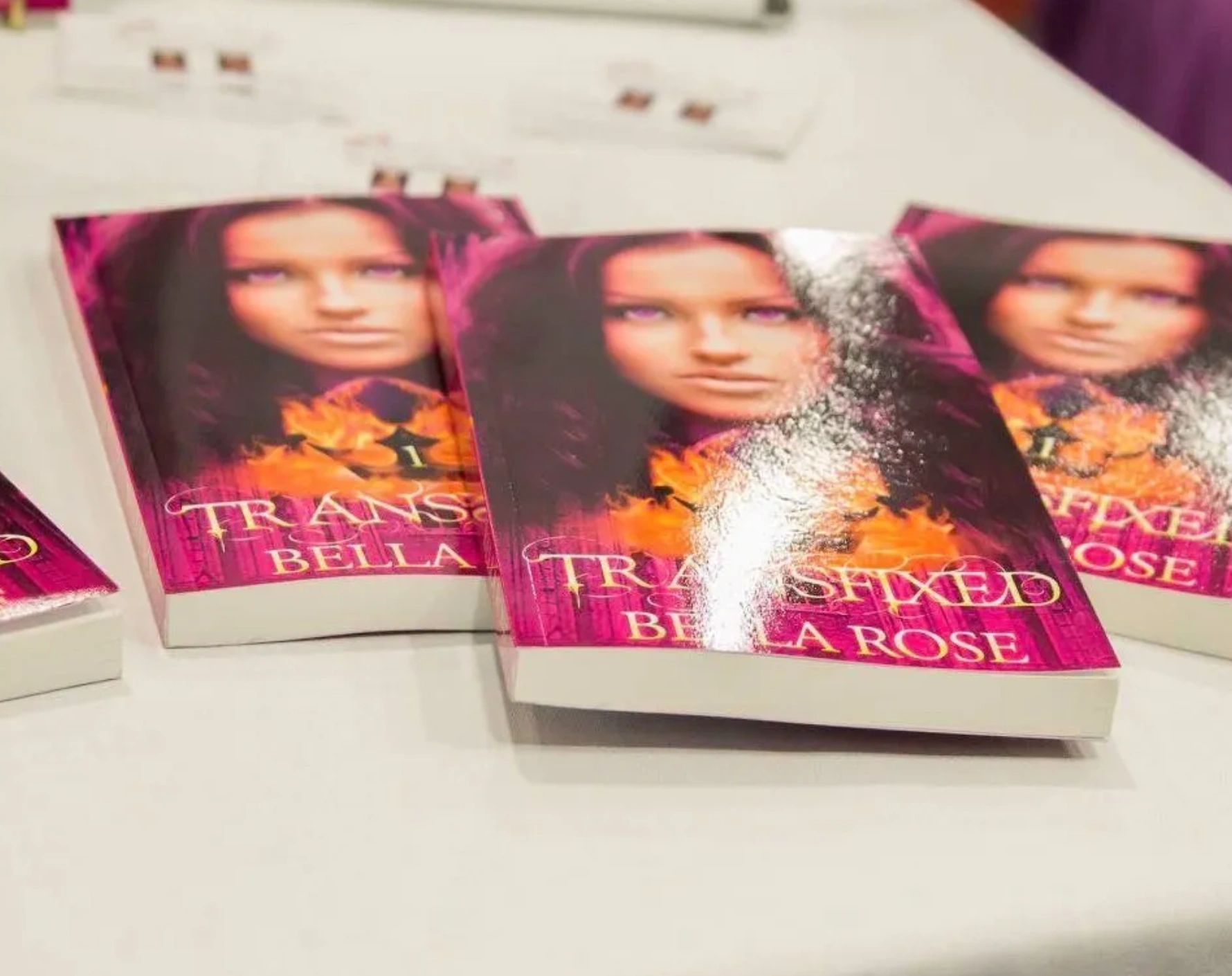 Stack of fantasy books titled 'Transfixed Bella Rose' with a fiery cover and a woman's face.
