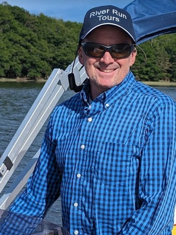 Man in blue checkered shirt and cap on a boat by the water.