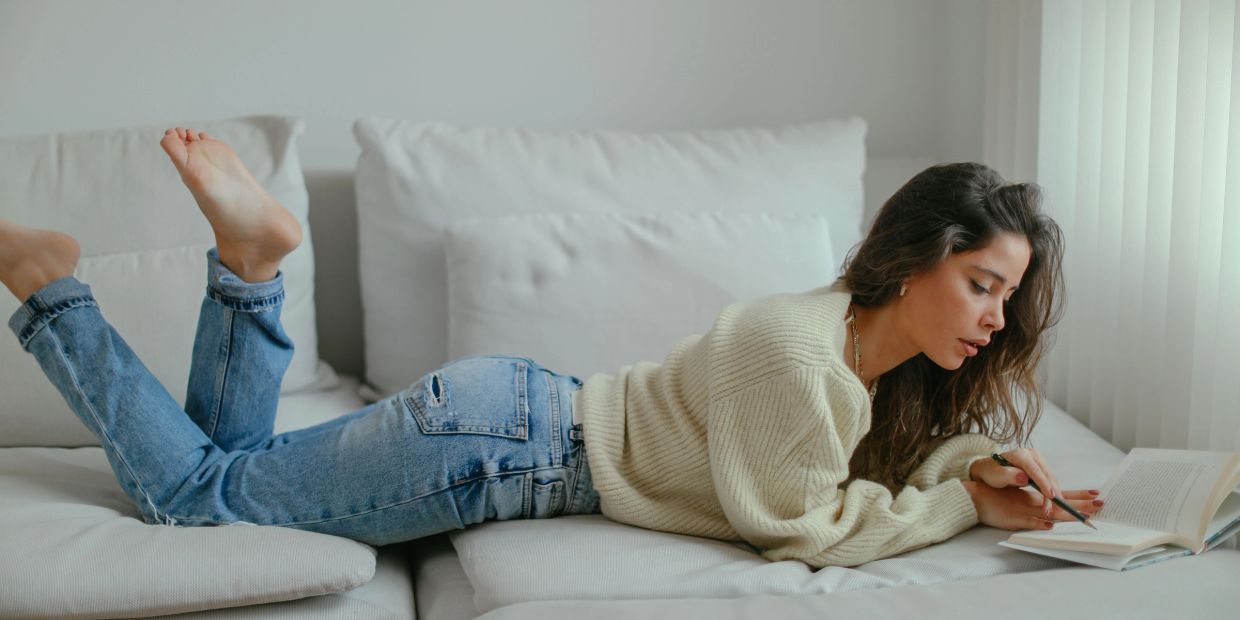 A woman in a cozy sweater and jeans lies on a sofa, writing in a book near a window.