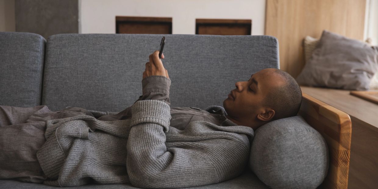 Person lying on a couch, looking at a phone in a cozy setting.