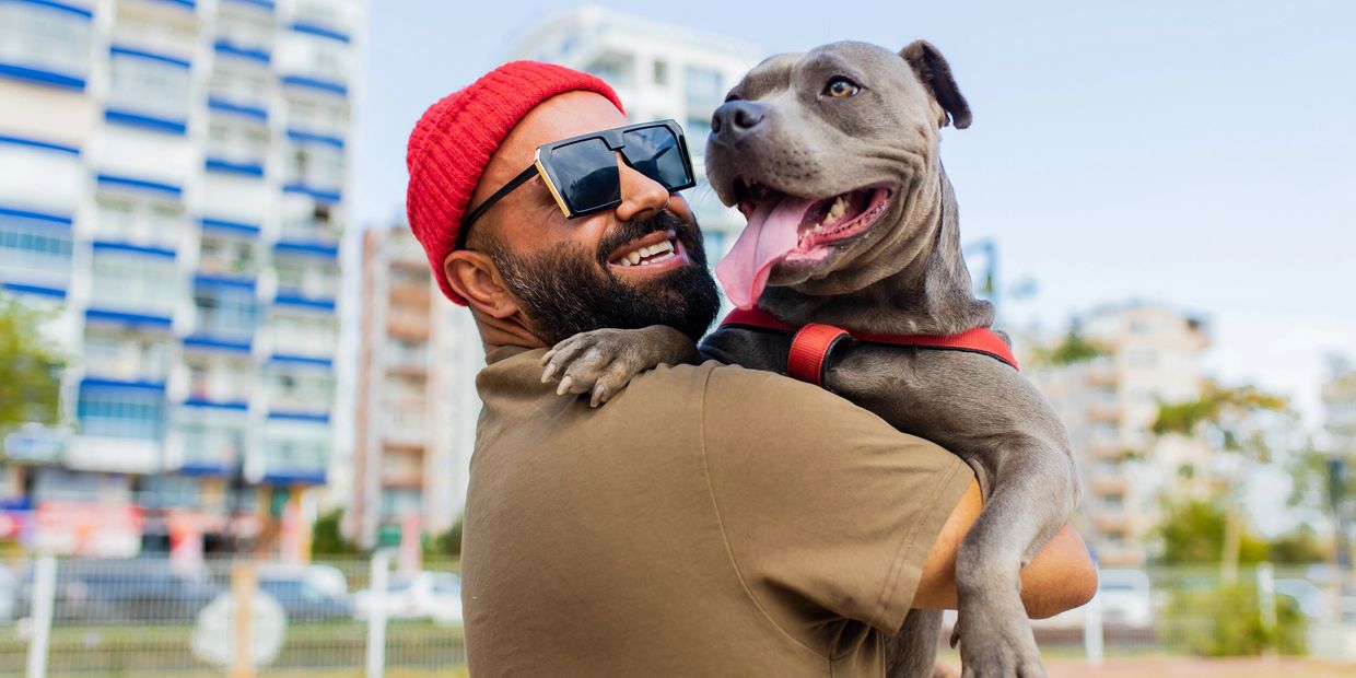 A man in a red hat with sunglasses giving his dog a hug