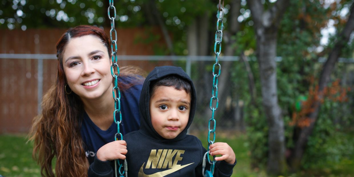 A woman smiles behind a young boy on a swing in a garden.