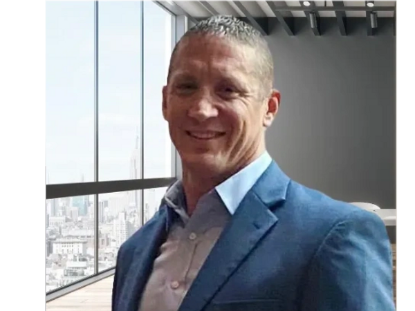 Man in a blue suit smiling in a modern office with city views.