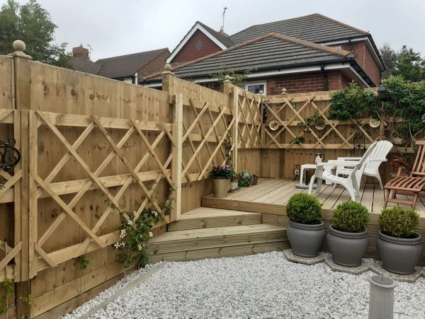 Cozy backyard with wooden deck, white chairs, potted plants, and decorative fence trellis.