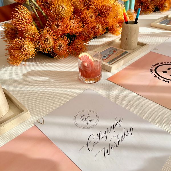 Elegant calligraphy workshop setup with vibrant orange flowers and refreshing drink.