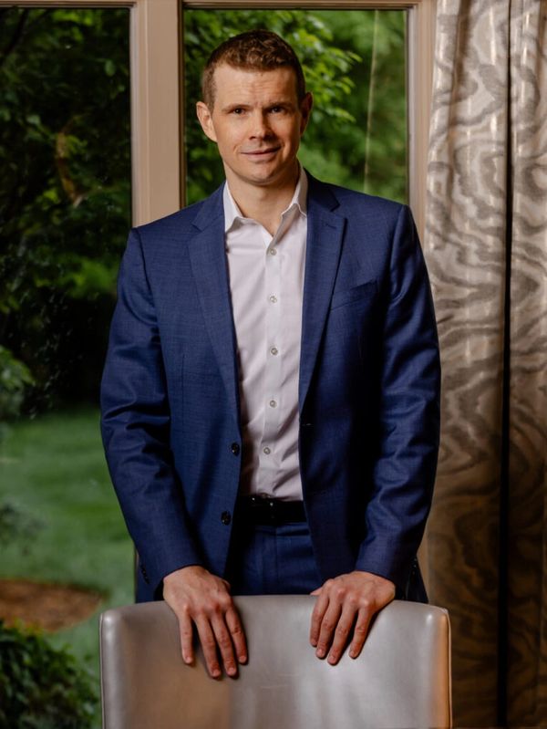 Man in a blue suit standing behind a chair indoors with greenery outside the window.