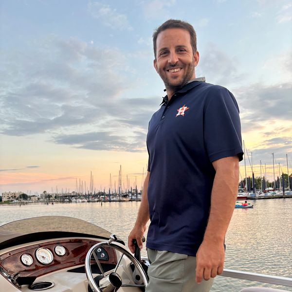Man steering a boat at sunset with a marina in the background.