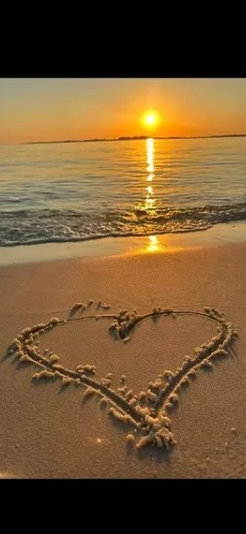 A heart drawn in the sand with the water and a sunset in the background
