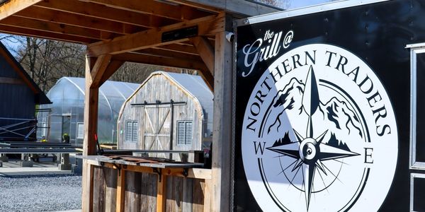 Outdoor grill stall at Northern Traders with rustic wooden sheds in background.
