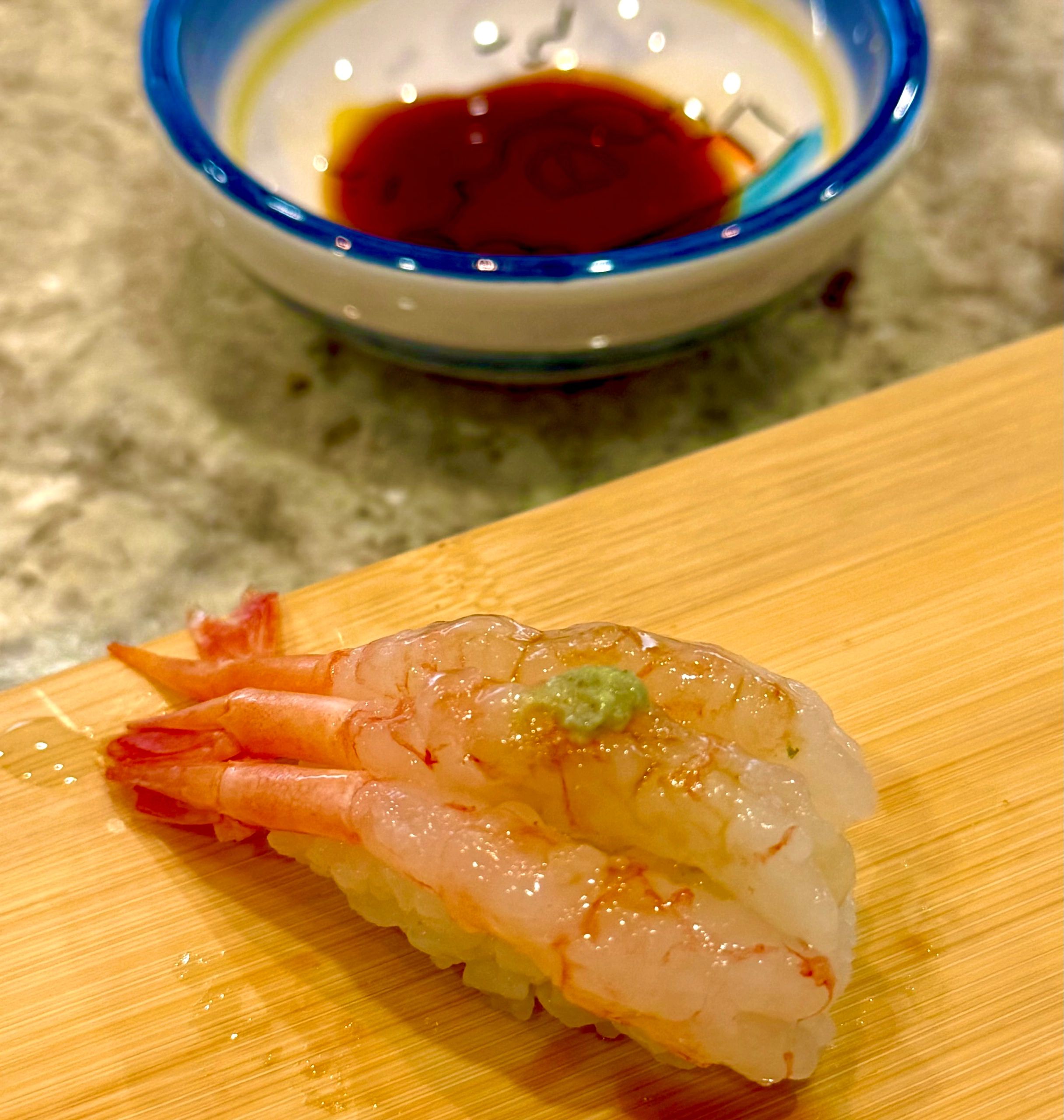 Fresh shrimp nigiri sushi with a dab of wasabi on top and soy sauce in the background.