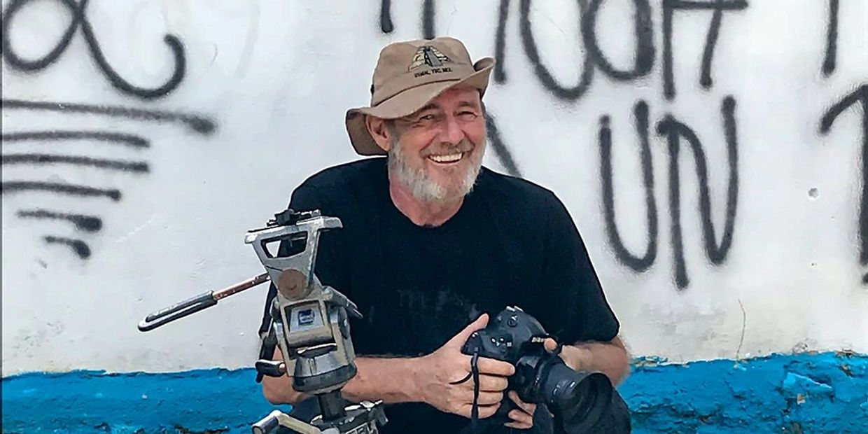 Michael A. Maurus sitting behind a tripod with a camera in his hands, smiling at the viewer.