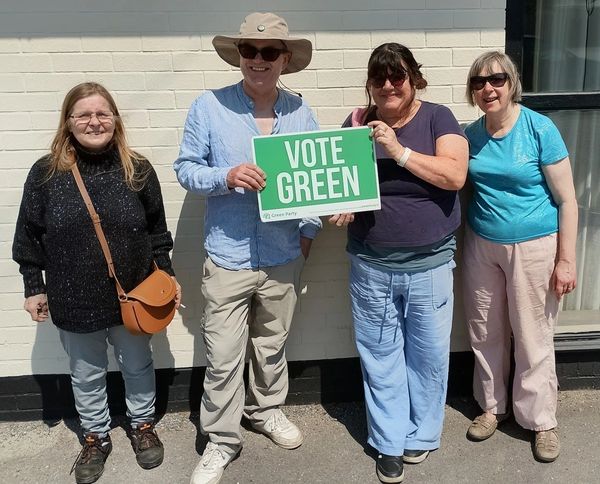Four people smiling, holding a 'Vote Green' sign outside on a sunny day.