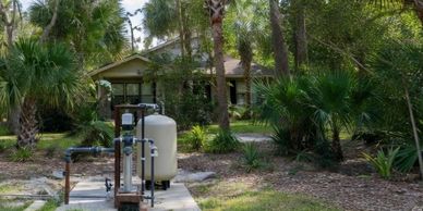 Water filtration system in a lush backyard with tropical plants and a house.