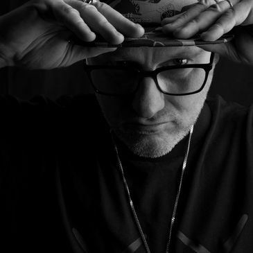 Man with glasses adjusting his cap, wearing rings and a pendant necklace.