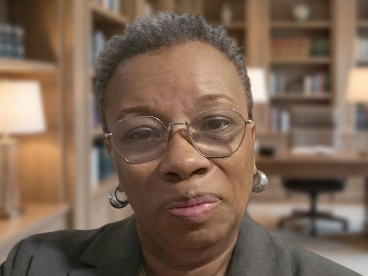 A serious older woman wearing glasses and earrings in a cozy study room.