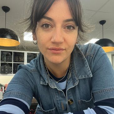 Young woman with short hair and hoop earrings wearing a denim jacket indoors.