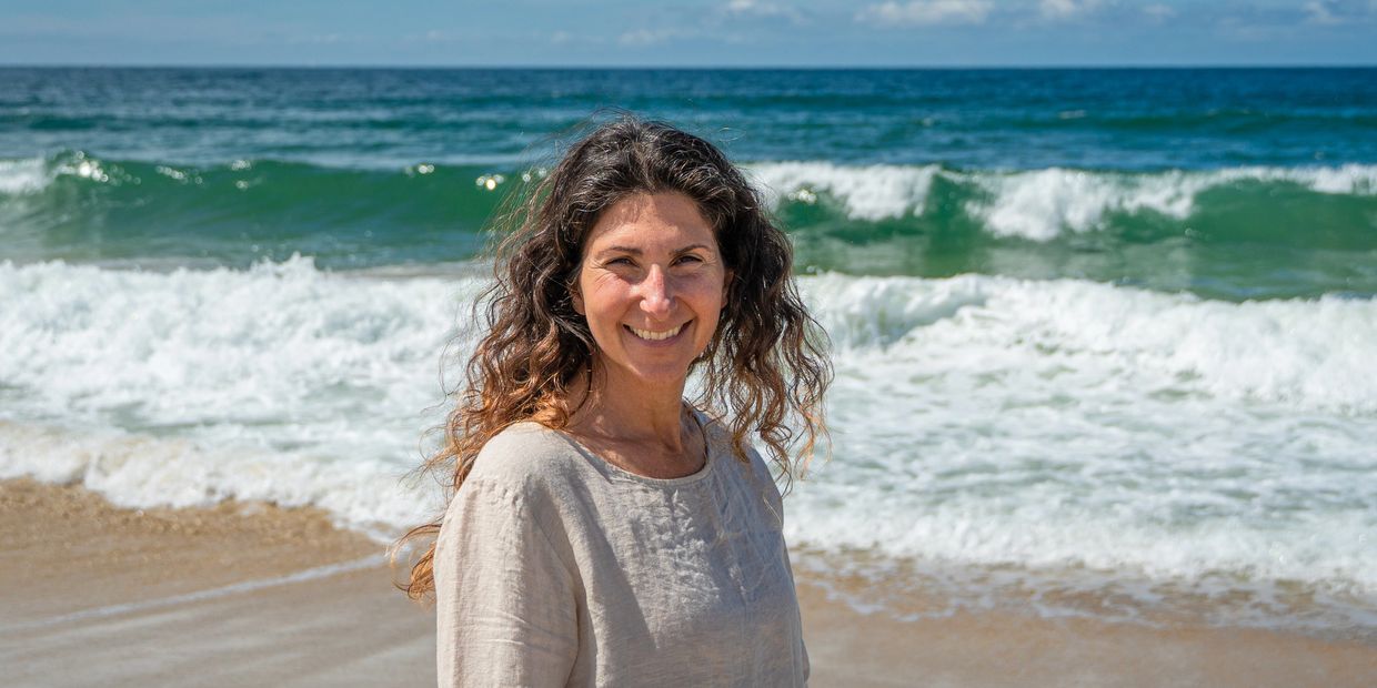 Smiling woman with curly hair stands on a sunny beach with ocean waves behind her.