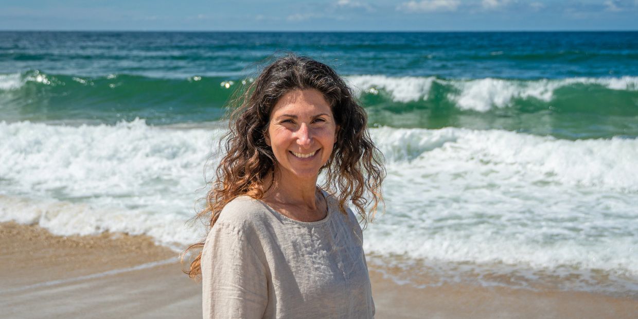 Smiling woman with curly hair stands on a sunny beach with ocean waves behind her.