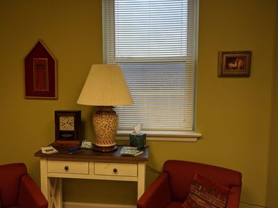 Cozy corner with lamp, clock, tissues, and red chairs near a window.