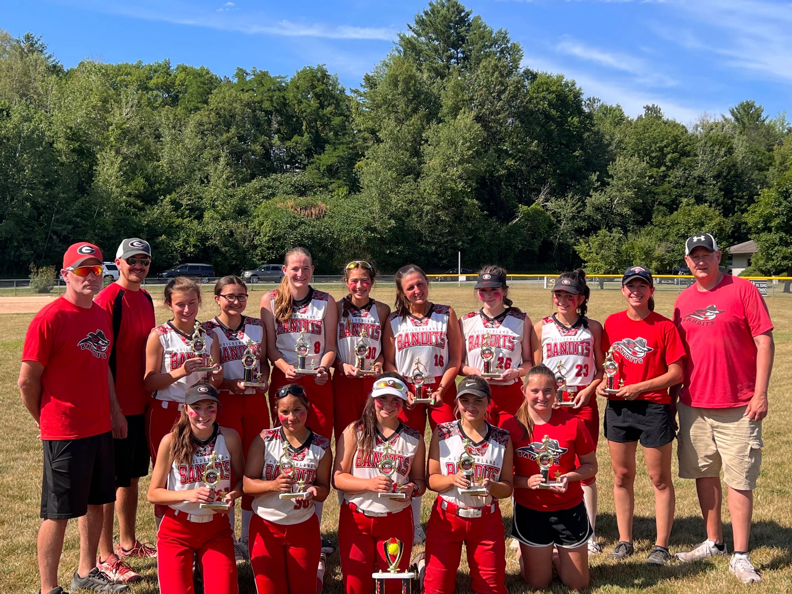 Youth softball team with coaches posing with trophies on a sunny day.