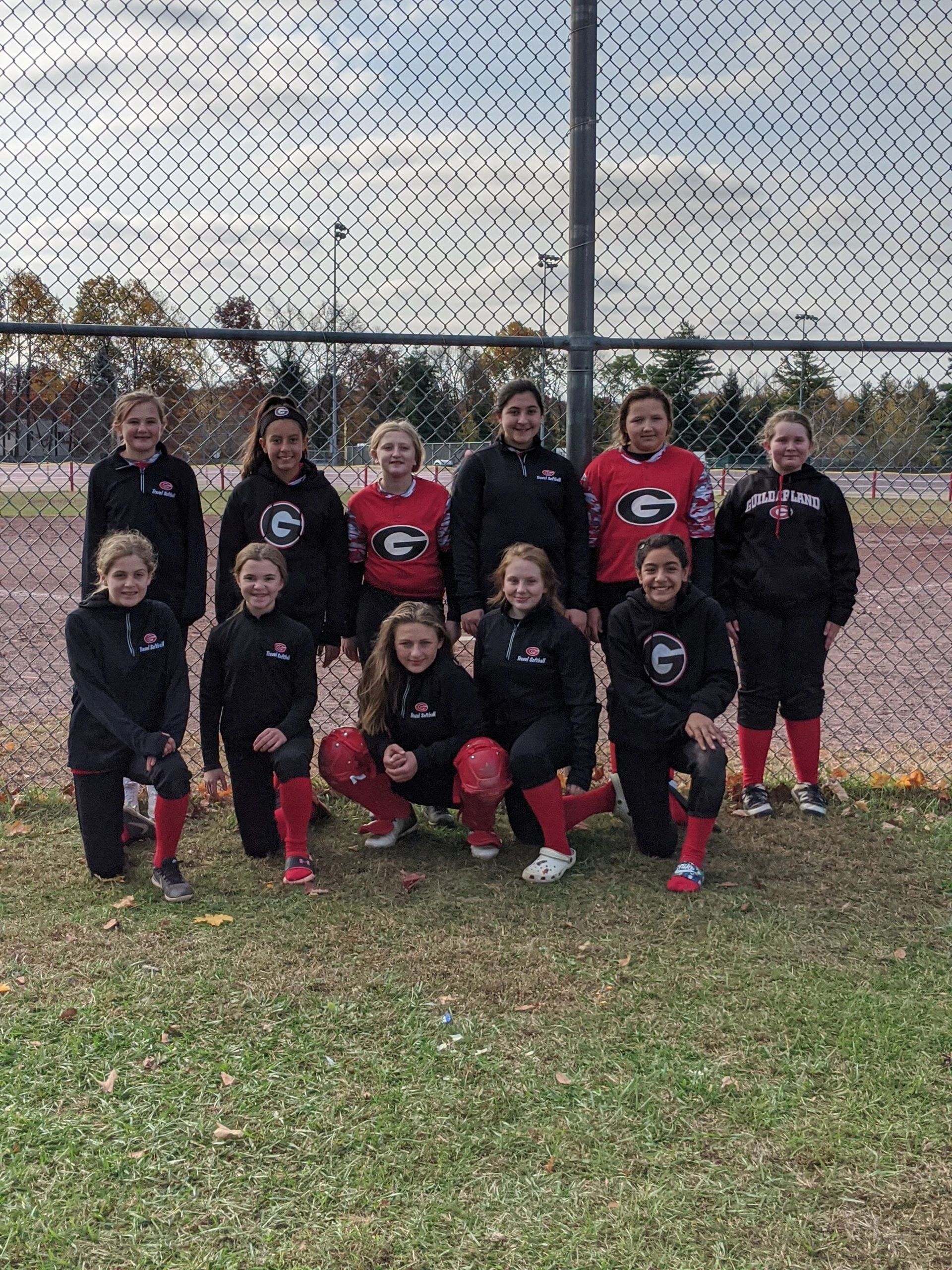 A youth softball team posing in front of a chain link fence on a field.