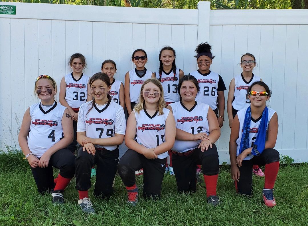 Youth softball team posing in white jerseys and black pants in front of a white fence.